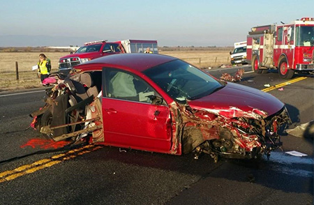 Red vehicle with damage to the front and back with a fire truck and ambulance in the background